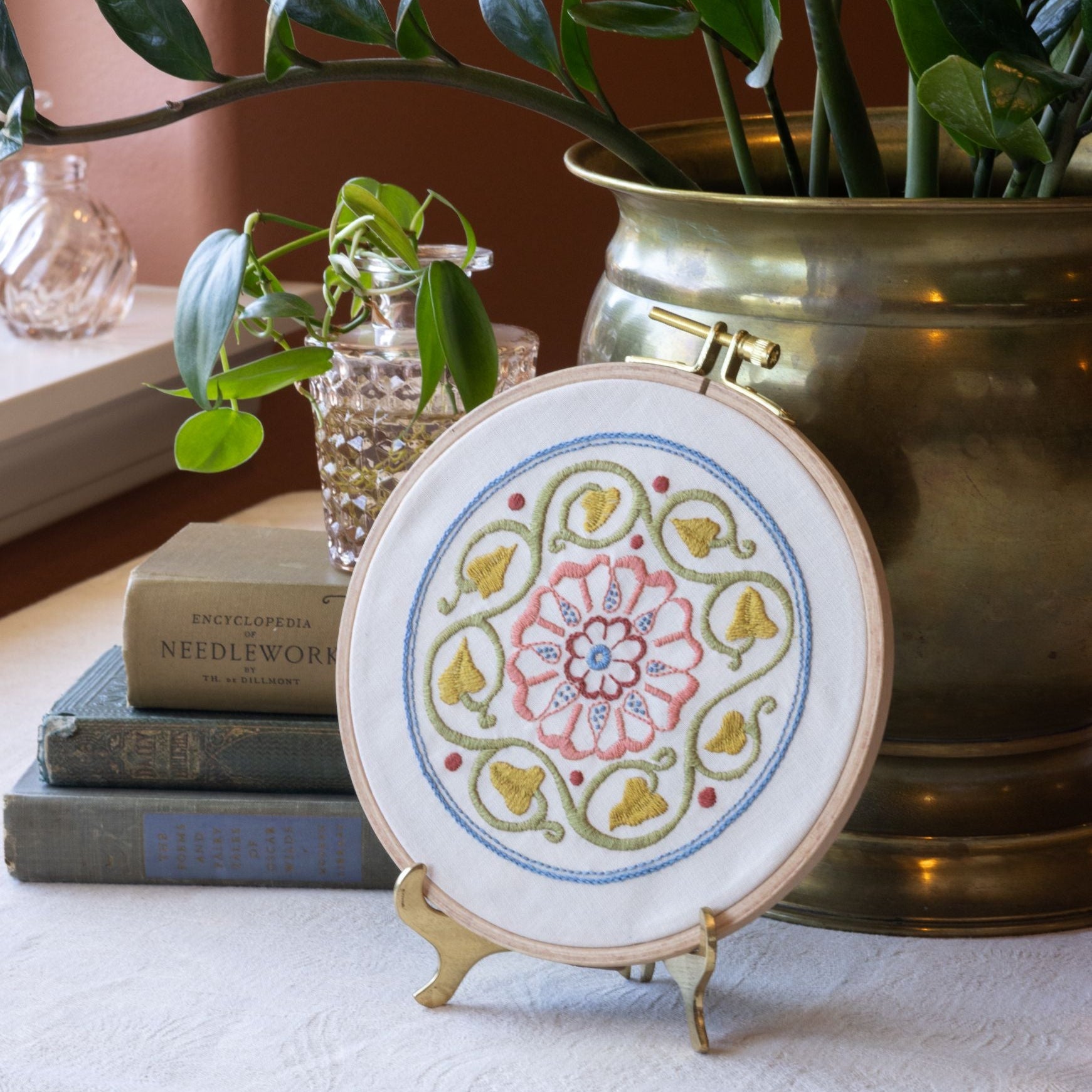 Embroidery hoop with floral design on a stand, surrounded by books and plants.
