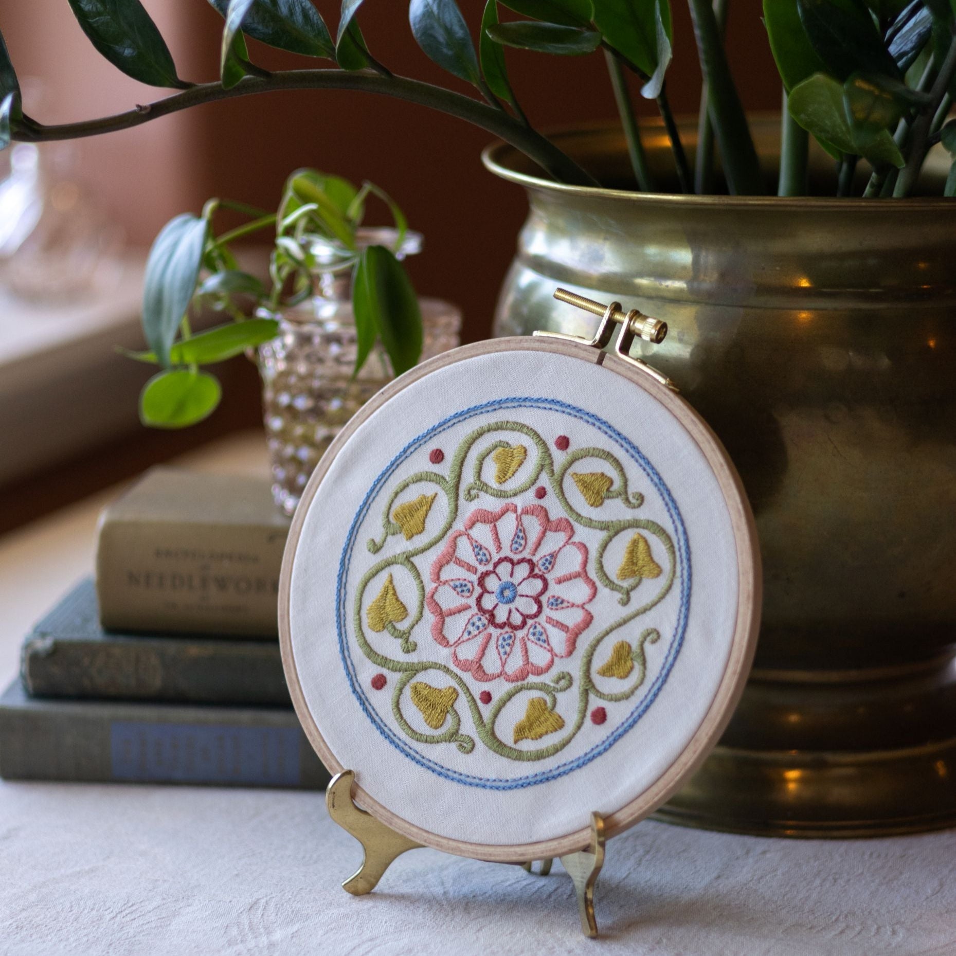 Embroidery hoop with floral design on a stand, placed on a surface with books and a plant in the background.