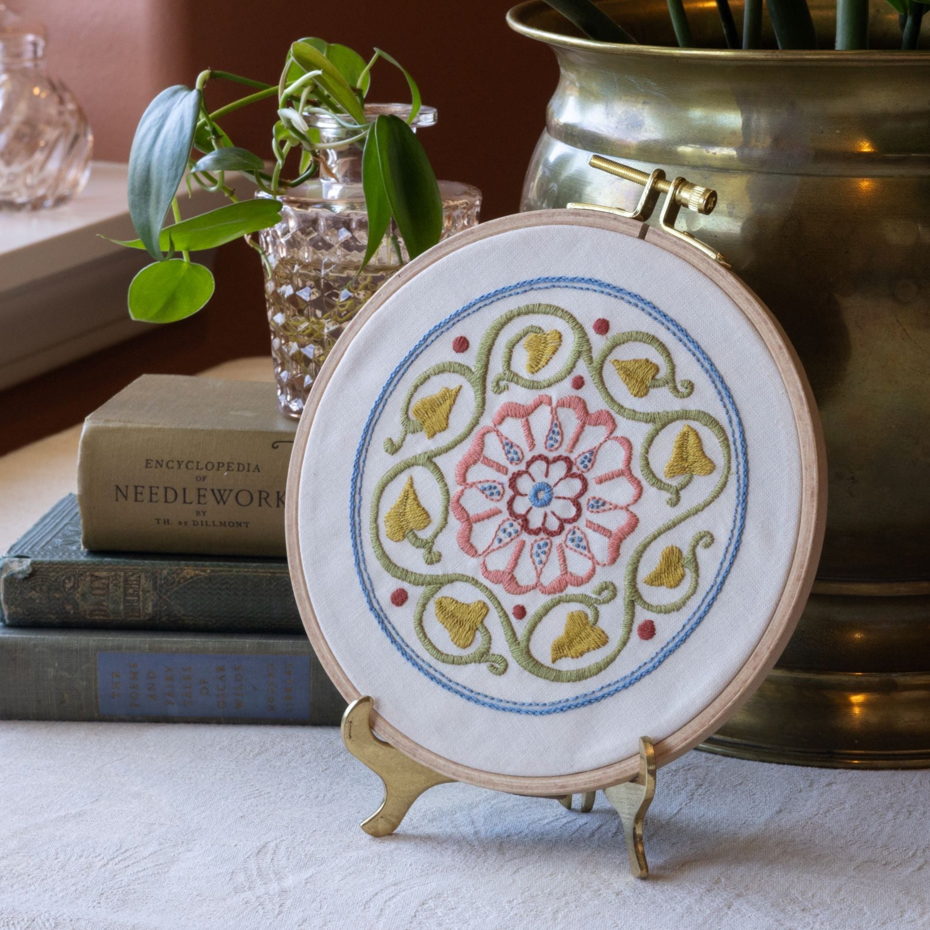 Embroidery hoop with floral design on a stand, books, and plant in the background