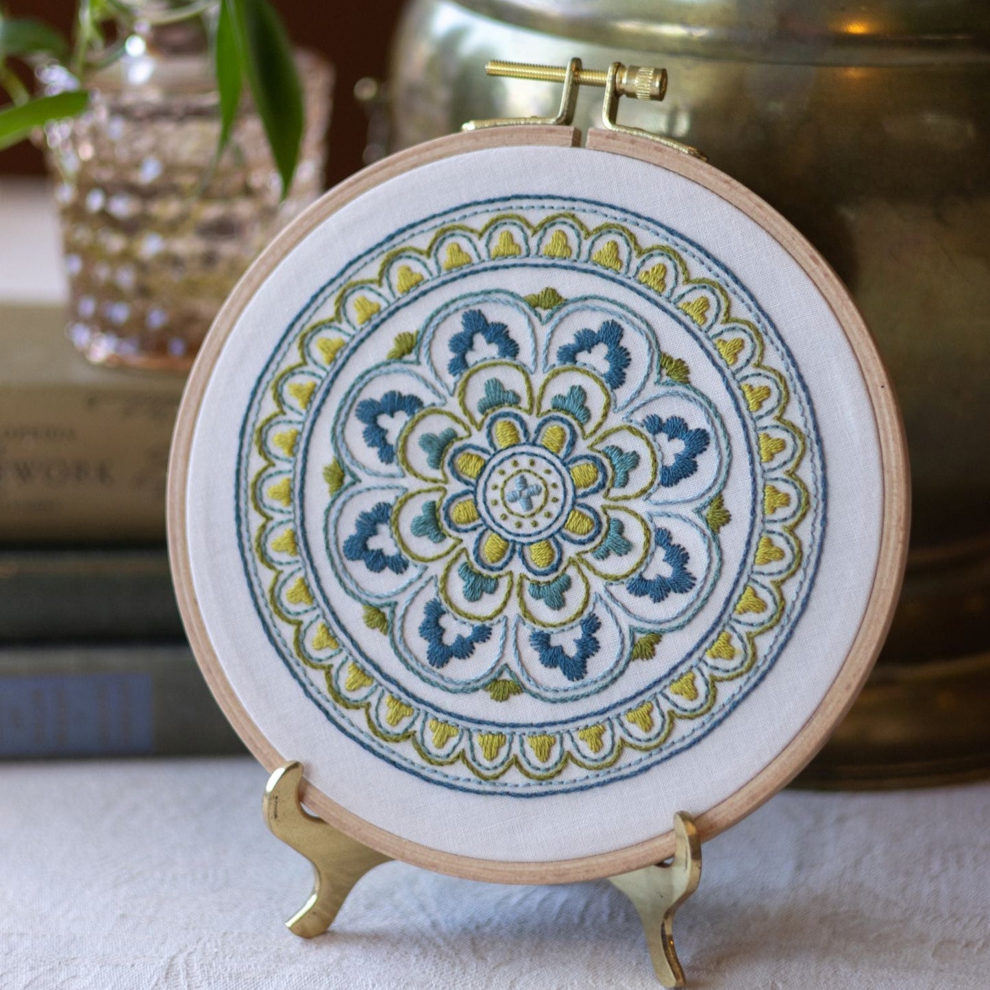 Embroidery hoop with a colorful mandala design on a stand, with a plant and books in the background.
