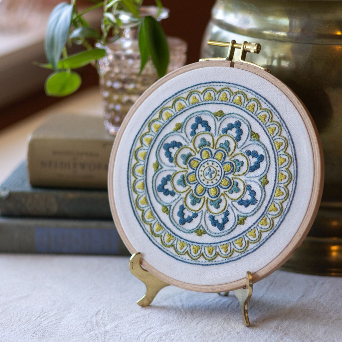 Embroidery hoop with mandala design on a stand, placed on a surface with books and plants in the background.