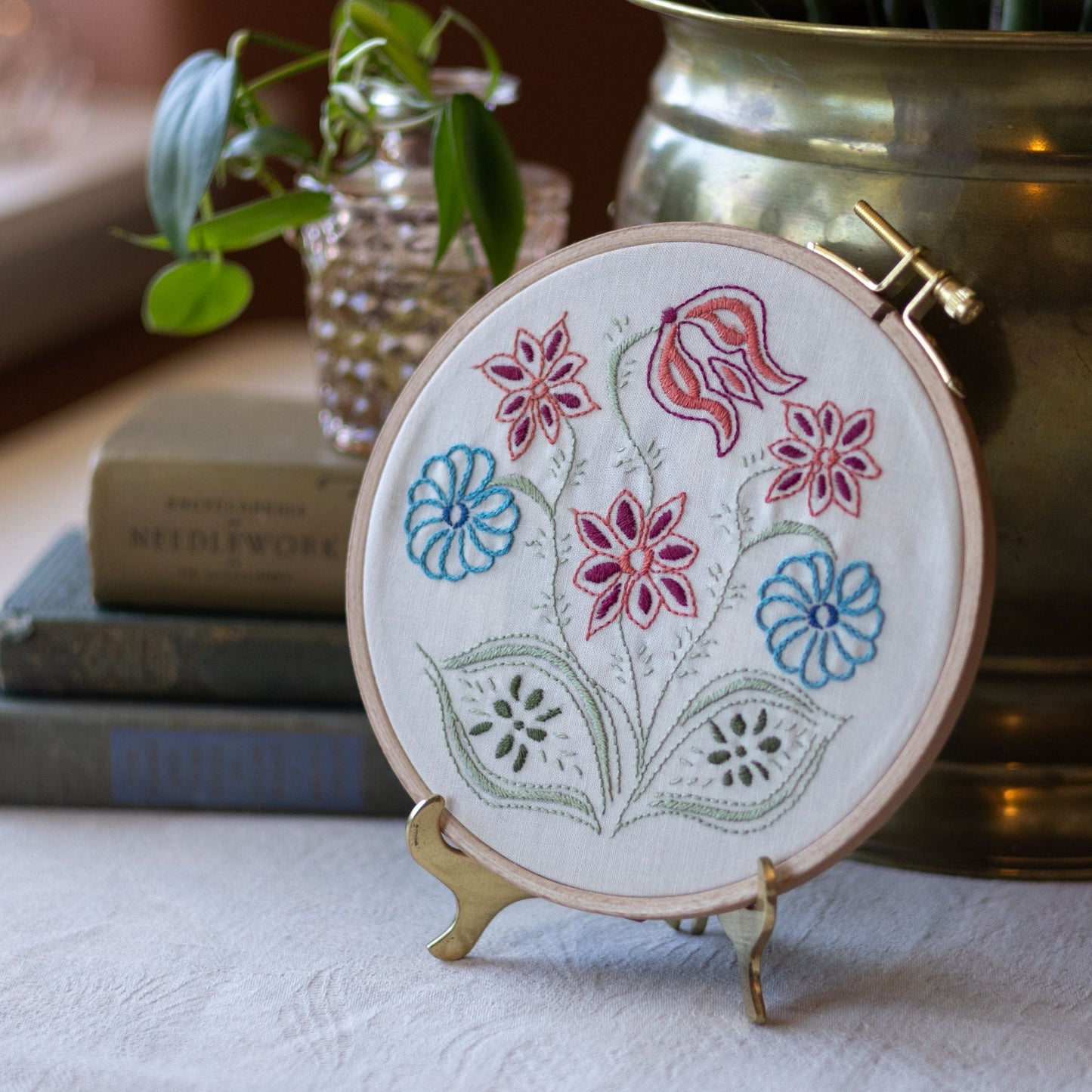 Embroidered hoop with floral design on a table with books and a plant in the background