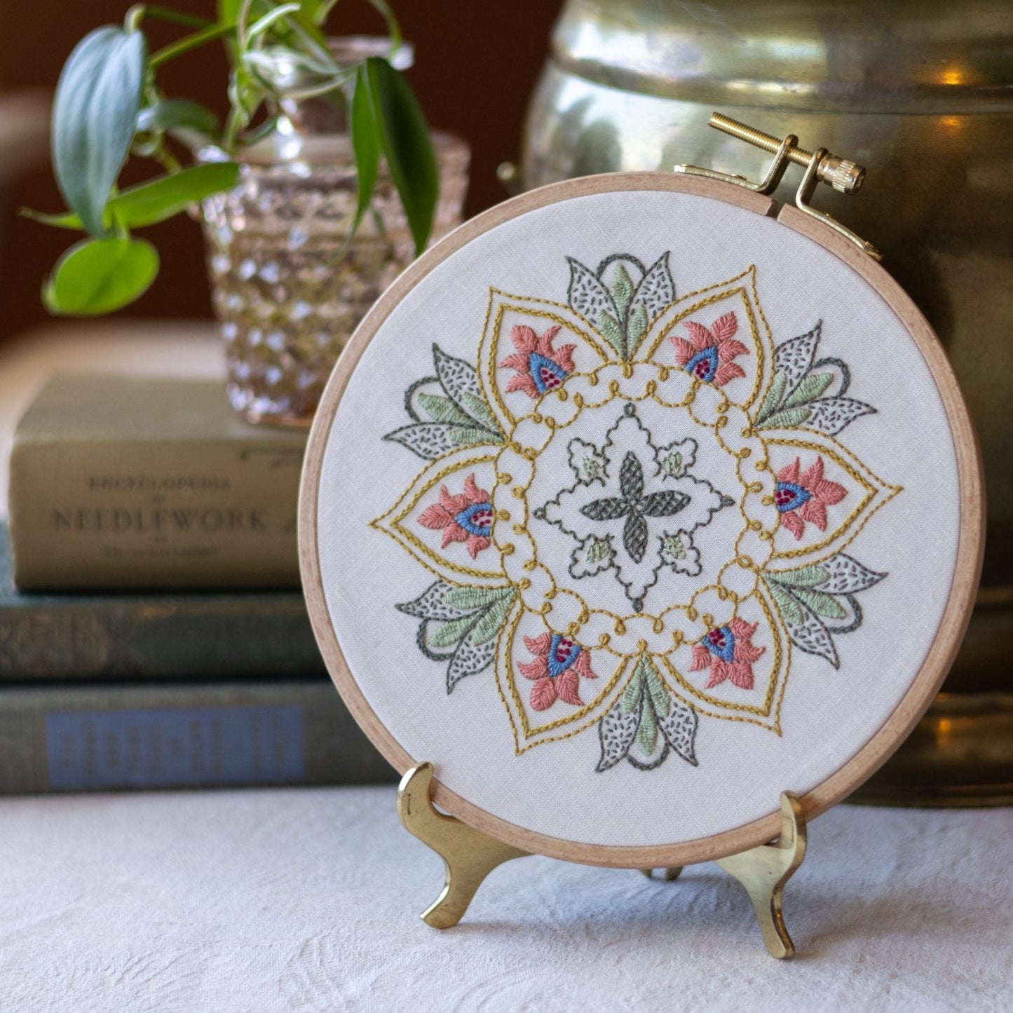 Embroidery hoop with floral design on a stand, placed on a surface with books and a plant in the background.