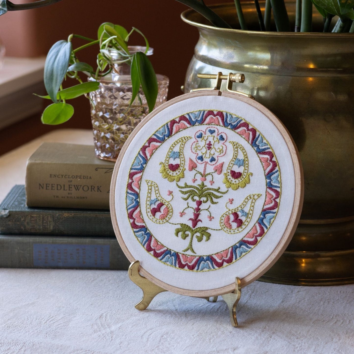 Embroidery hoop with floral design on a stand, placed on a surface with books and a plant in the background.