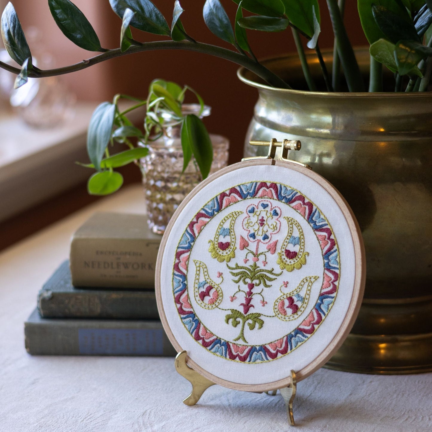 Decorative embroidery hoop with floral design on a stand, surrounded by books and plants.