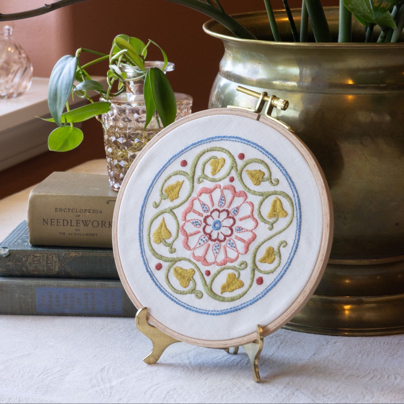 Embroidery hoop with floral design on a stand, surrounded by books and plants.