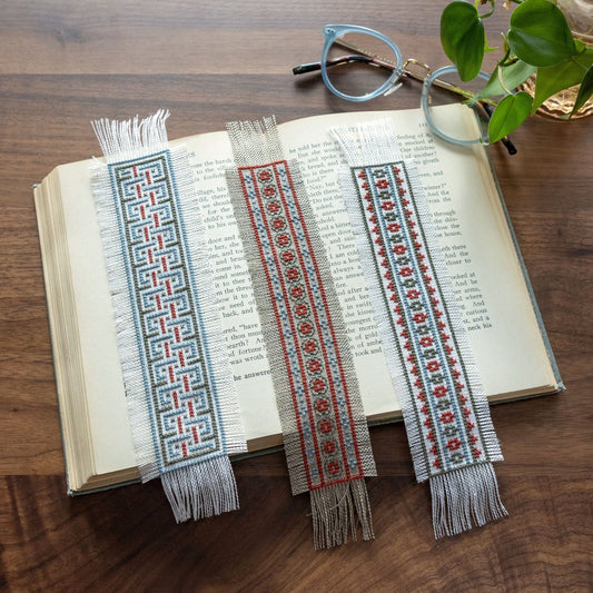 Three decorative cross stitch bookmarks on an open book with a wooden surface and glasses in the background.