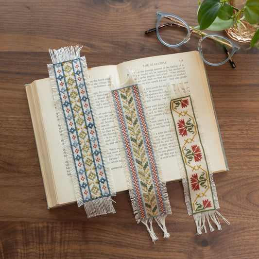 Three decorative bookmarks on an open book with glasses and a plant in the background.