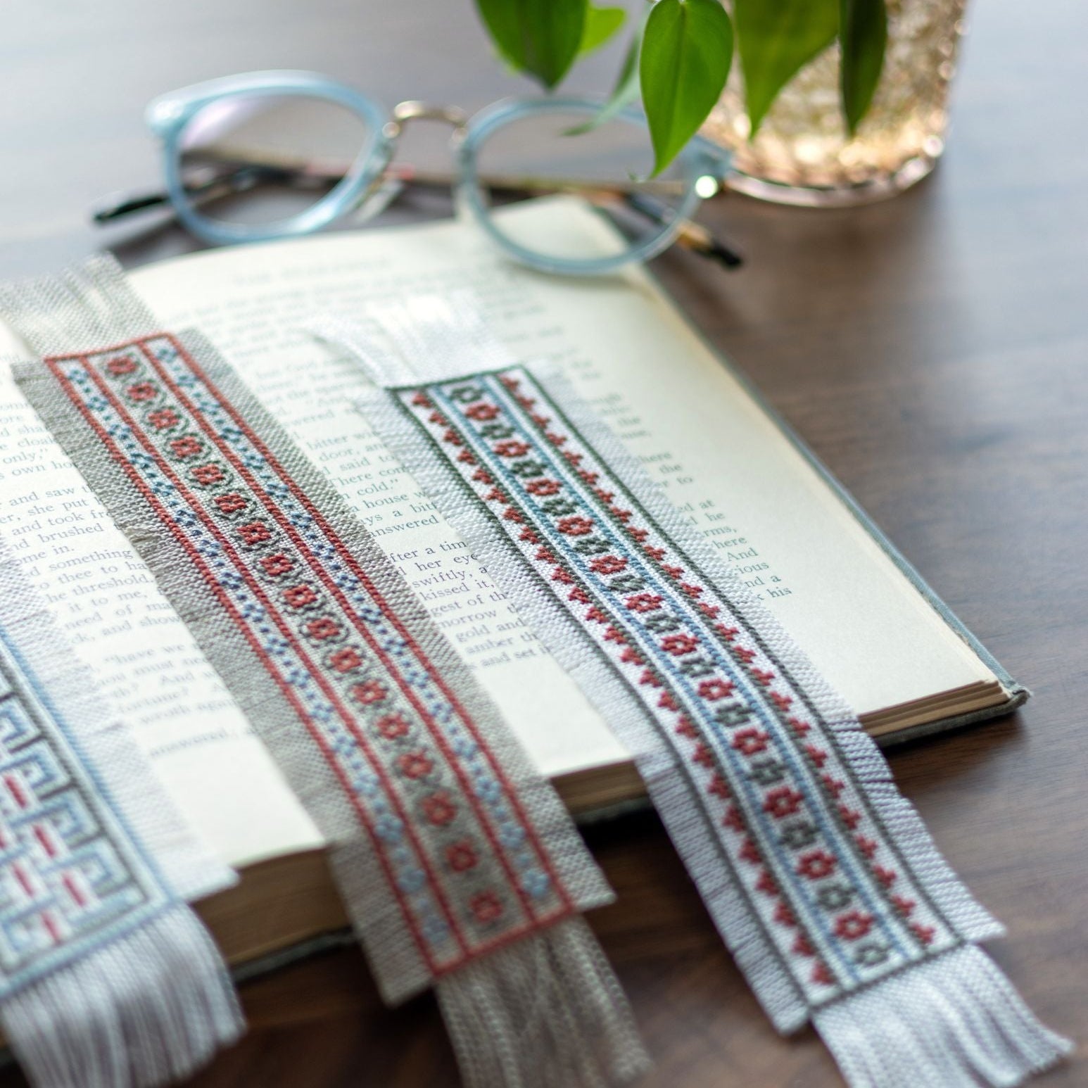 Decorative cross stitch bookmarks with intricate patterns on an open book, with a plant and glasses in the background.