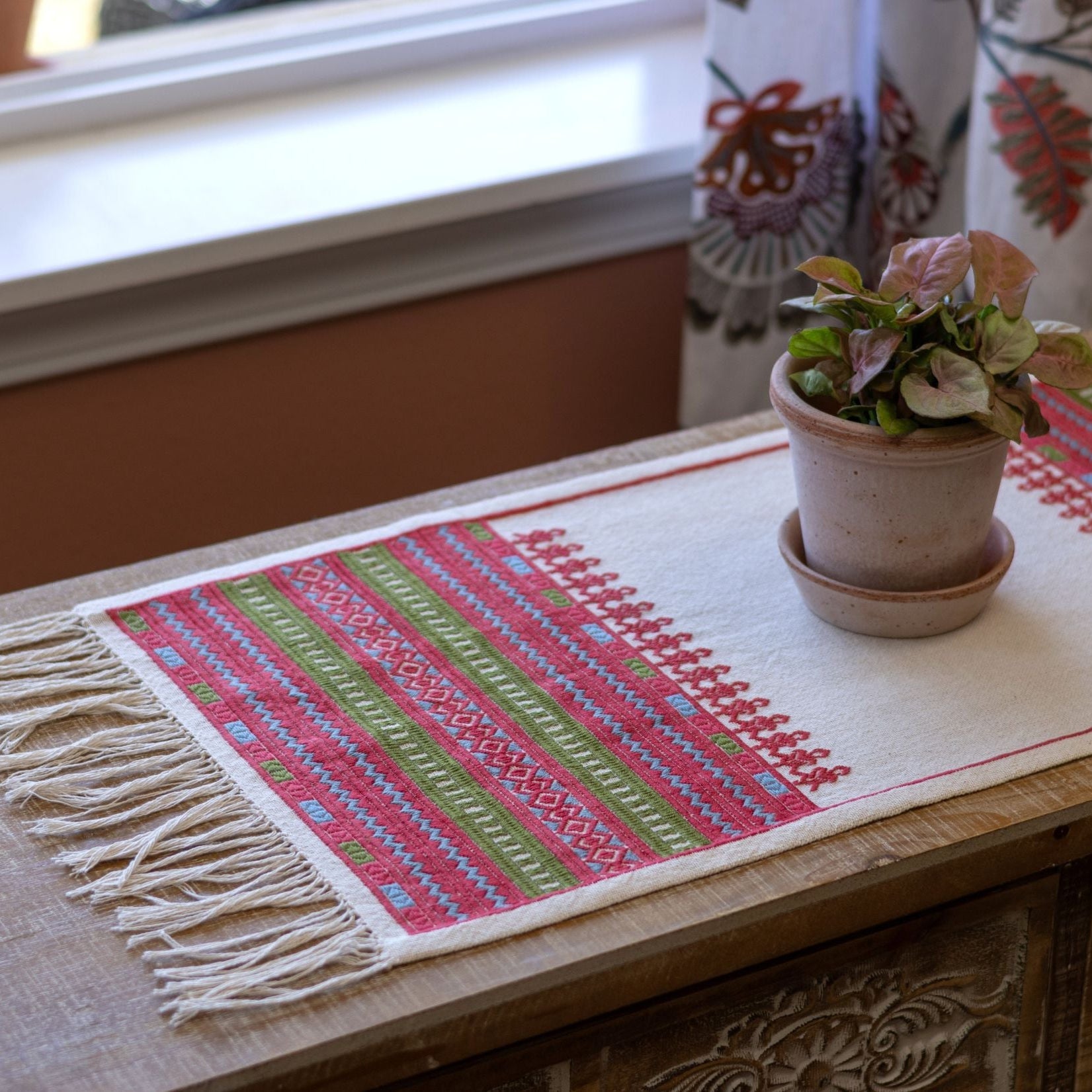 A Greek folk embroidery design with the atzolati satin stitch technique in red, green, and blue with knotted fringe ends on table with plant