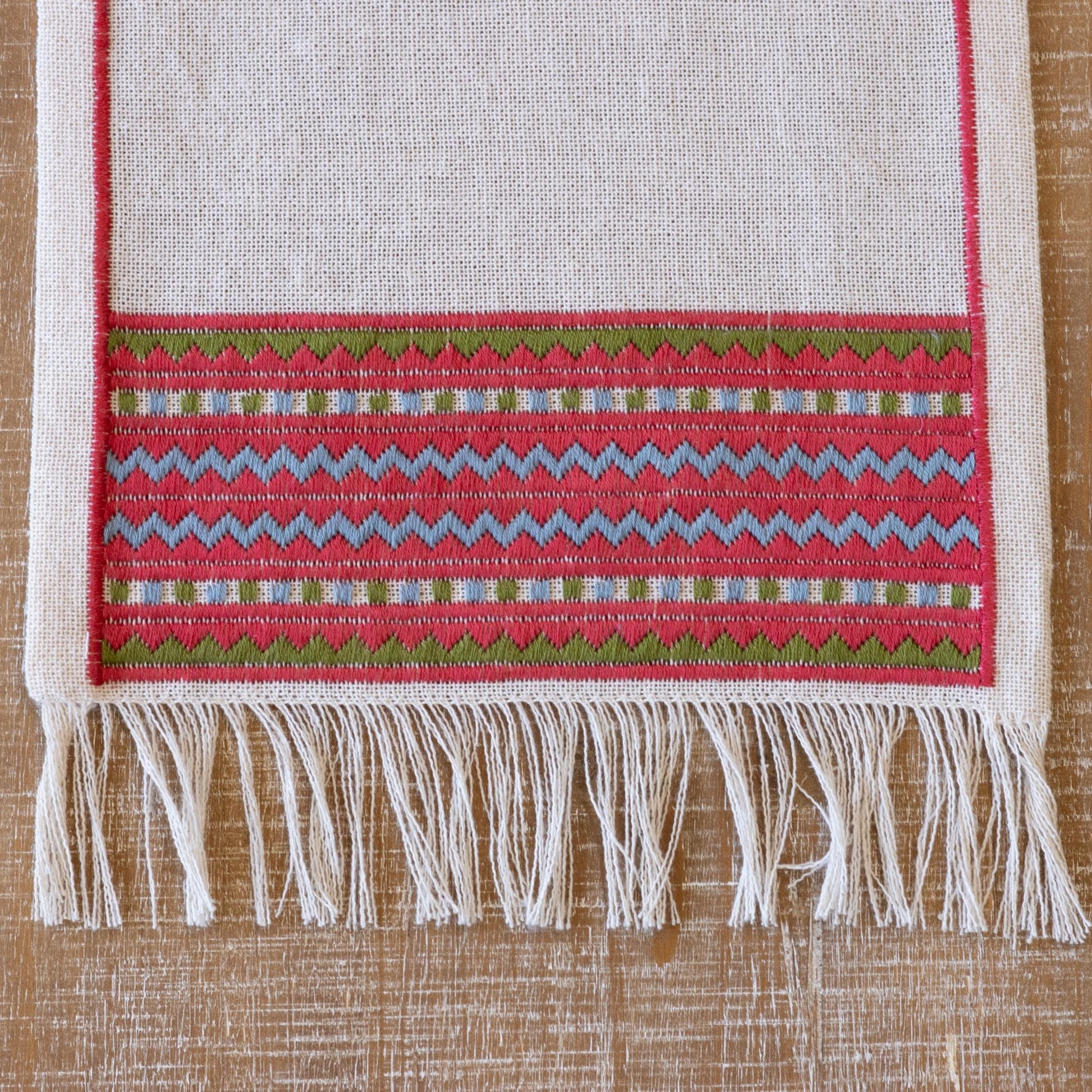 A close-up of an atzolati cross stitch folk embroidery textile with fringe, featuring a red border and geometric stripes in light blue and green, styled on a wooden table.
