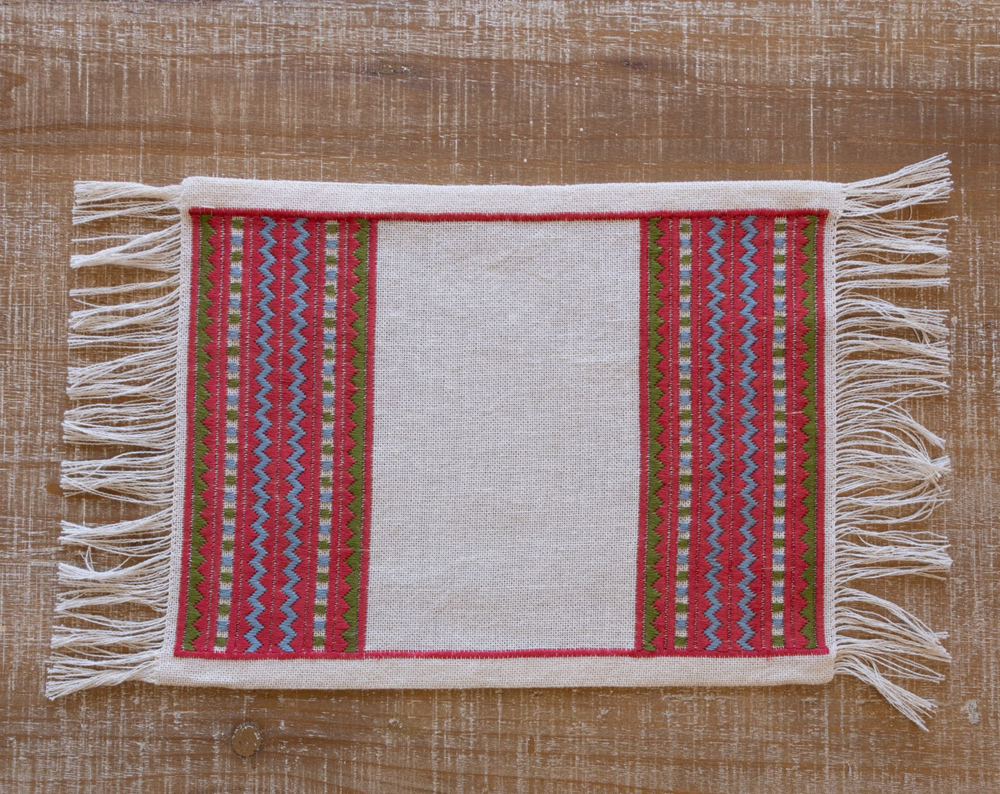 A close-up of an atzolati cross stitch folk embroidery textile with fringe, featuring a red border and geometric stripes in light blue and green, styled on a wooden table.