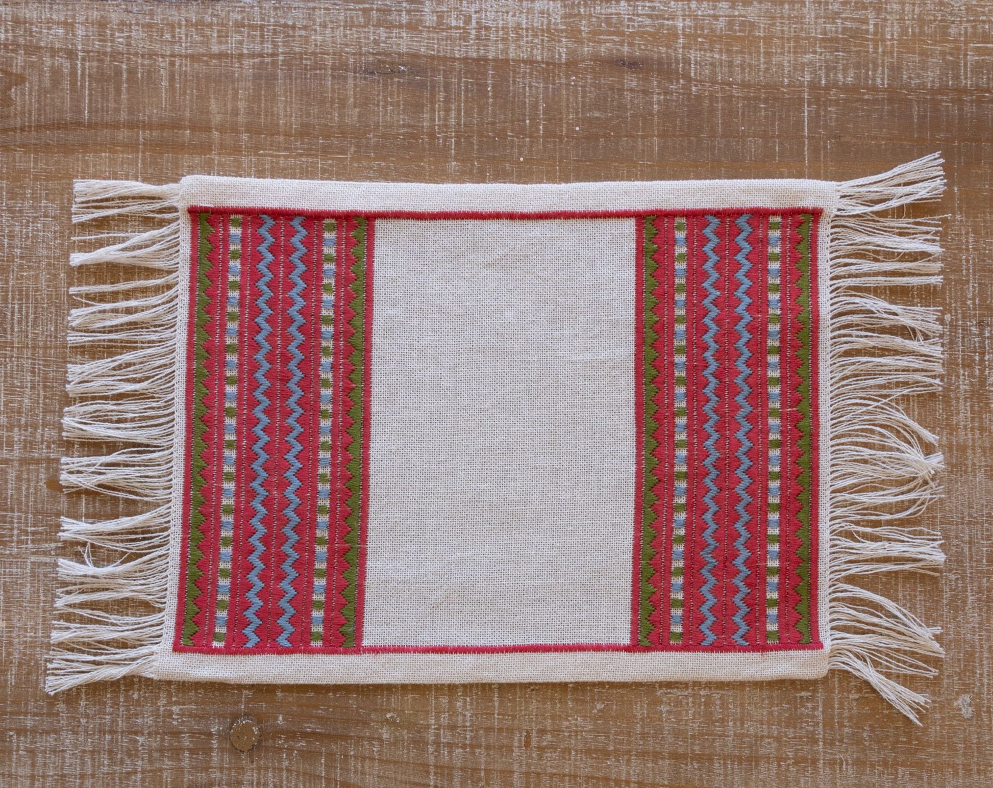A close-up of an atzolati cross stitch folk embroidery textile with fringe, featuring a red border and geometric stripes in light blue and green, styled on a wooden table.