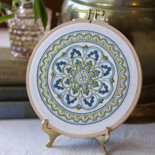 Embroidery hoop with a colorful mandala design on a stand, with a plant and books in the background.