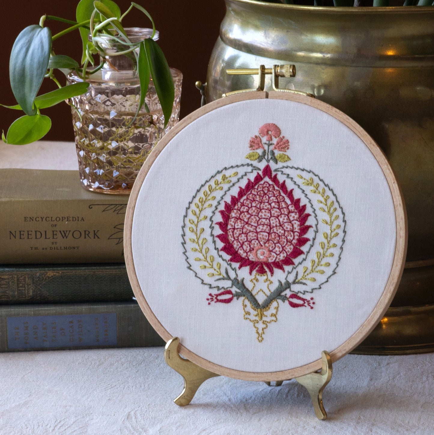 Embroidery hoop with floral design on a stand, books, and plant in the background