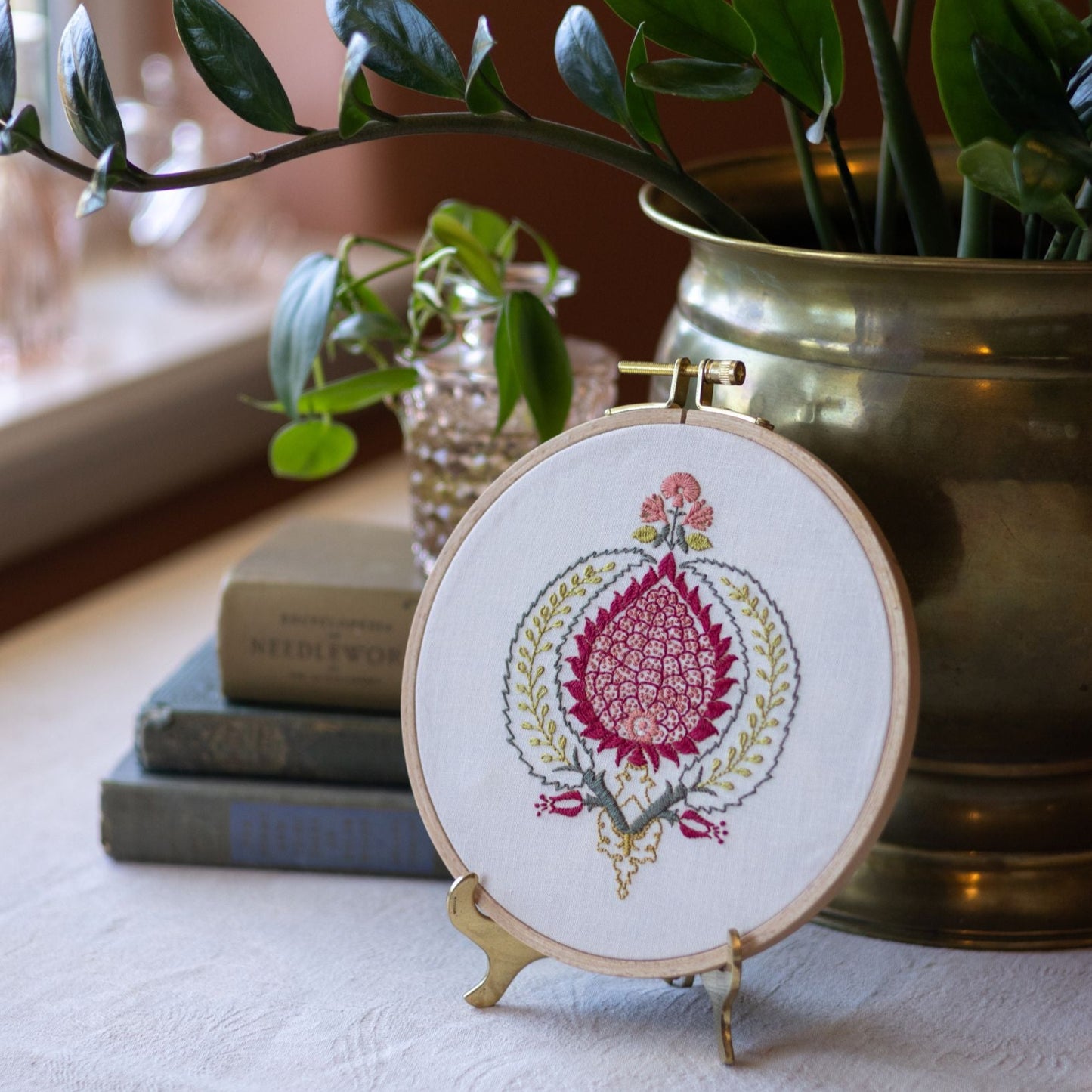 Embroidered hoop with floral design on a stand, placed on a surface with books and plants in the background.