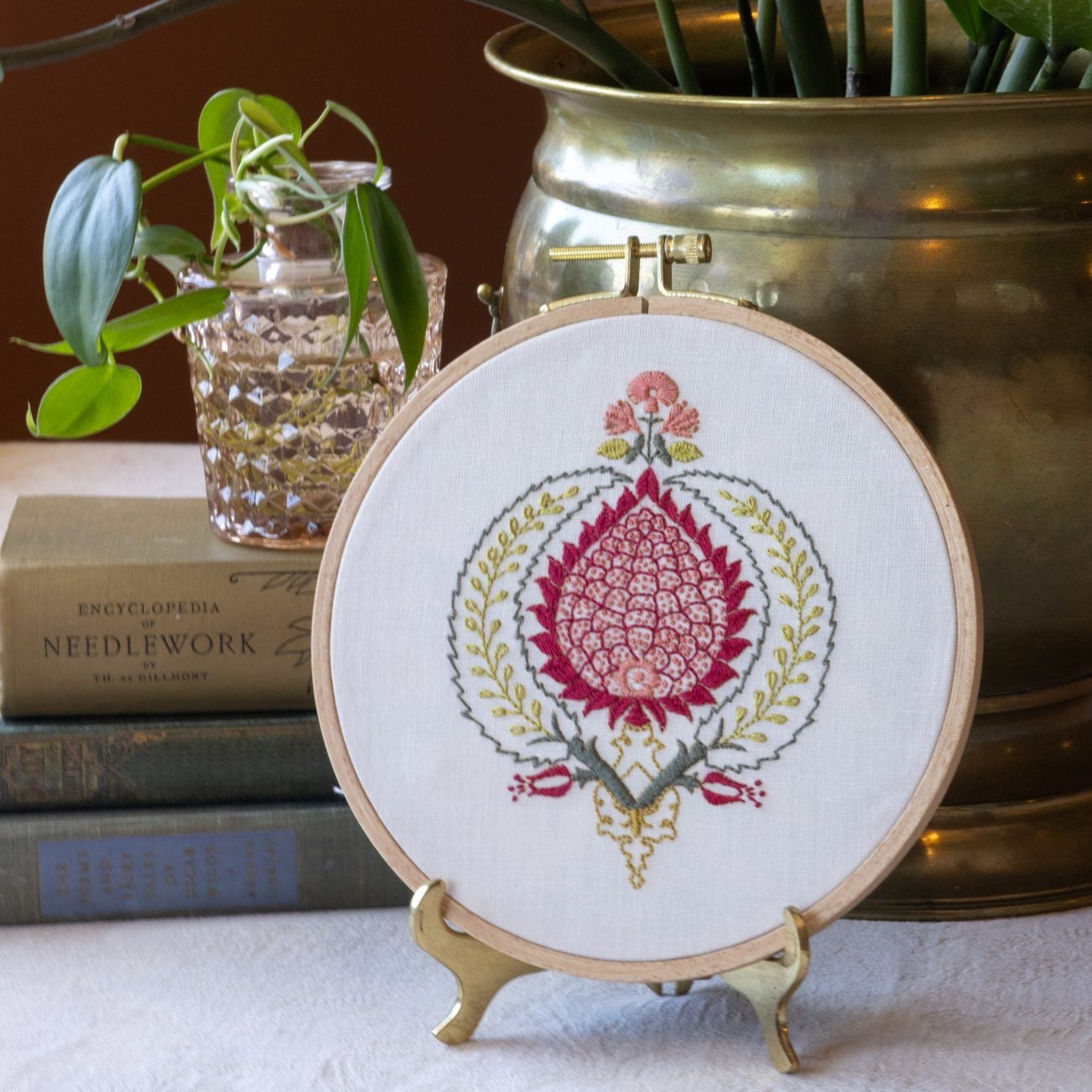 Embroidery hoop with floral design on a stand, placed in front of a plant and books.