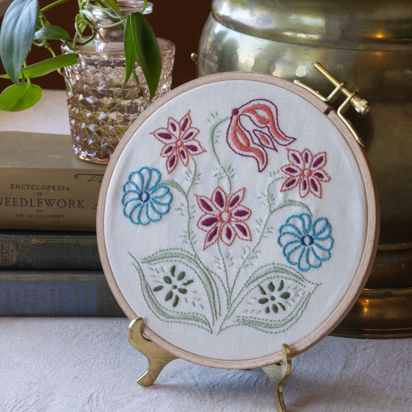 Embroidery hoop with floral design on a stand, books, and plant in the background