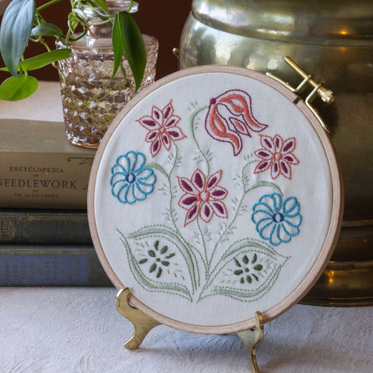 Embroidery hoop with floral design on a stand, books, and plant in the background
