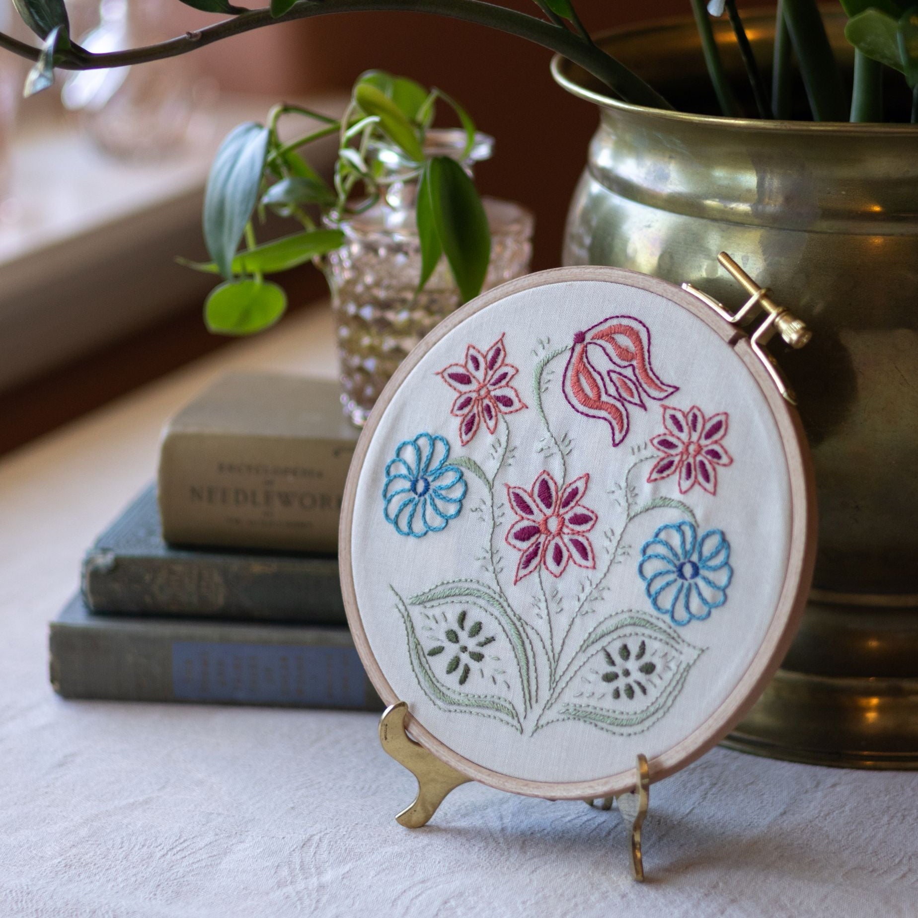 Embroidered hoop with floral design on a stand, surrounded by books and a plant.