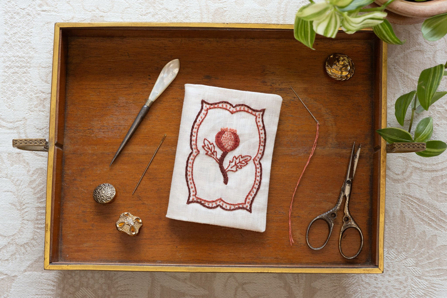 Wooden tray with embroidery tools and a needle book with pomegranate design on a light wooden surface.
