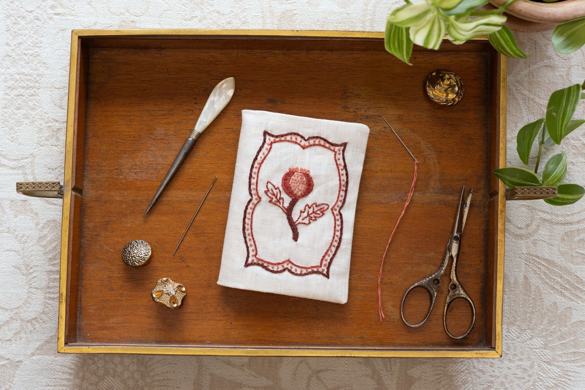 Wooden tray with embroidery tools and a needle book with pomegranate design on a light wooden surface.