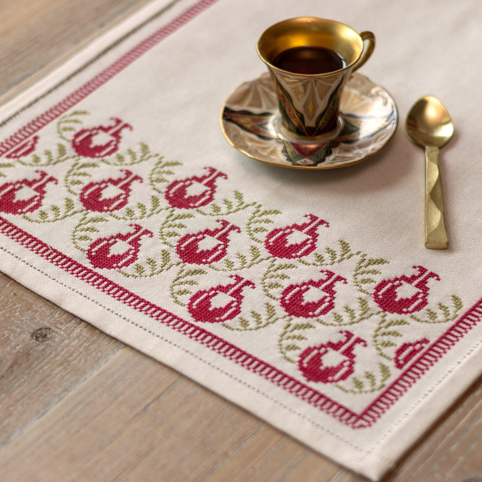 cross stitch table runner with pomegranate folk embroidery motif on wooden table with cup and spoon