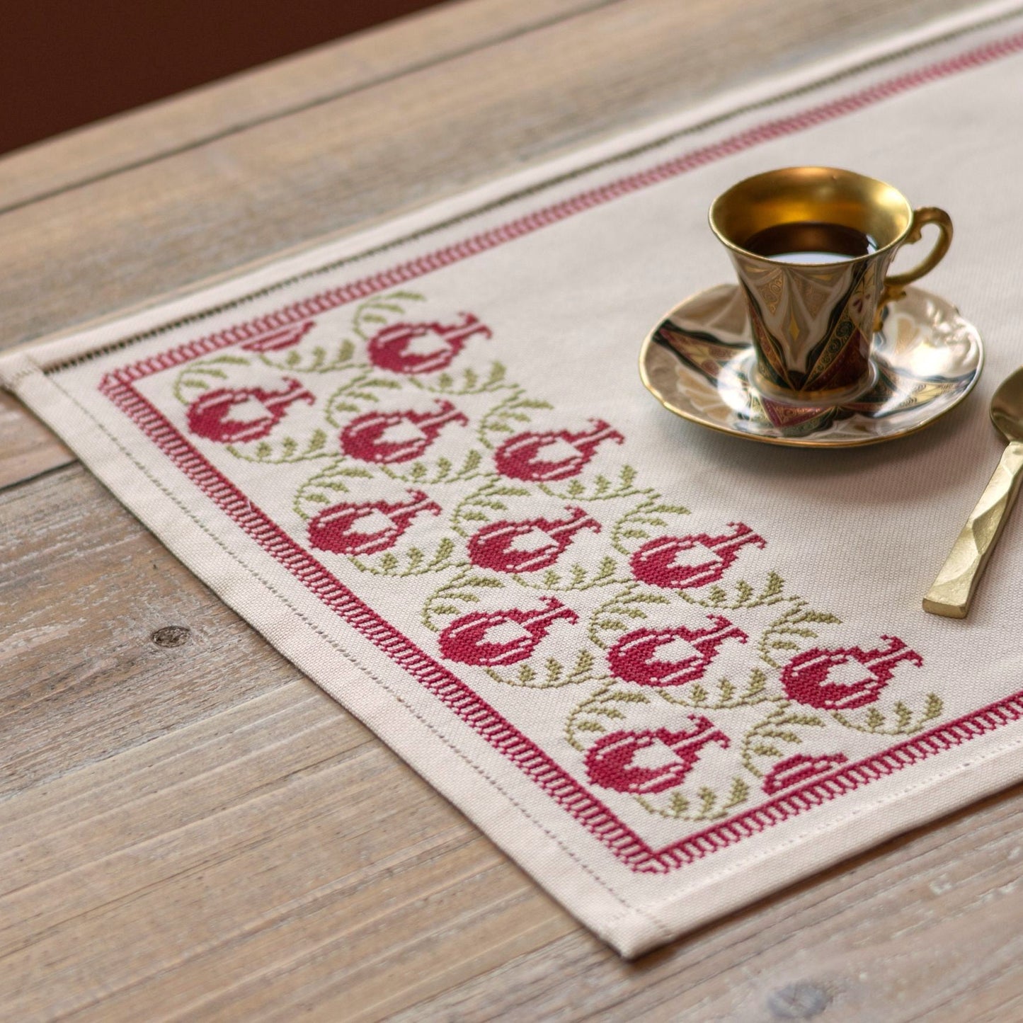 cross stitch table runner with pomegranate folk embroidery motif on wooden table with cup and spoon