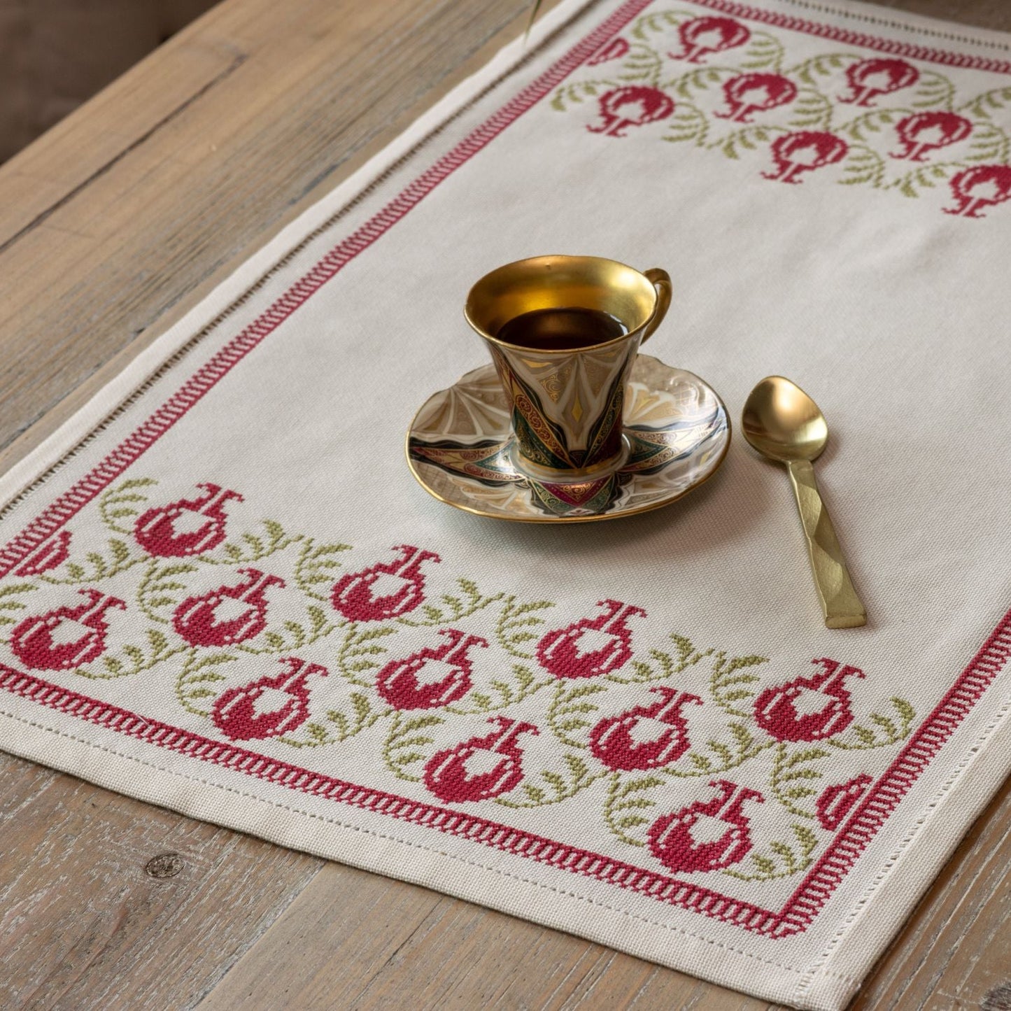 cross stitch table runner with pomegranate folk embroidery motif on wooden table with cup and spoon