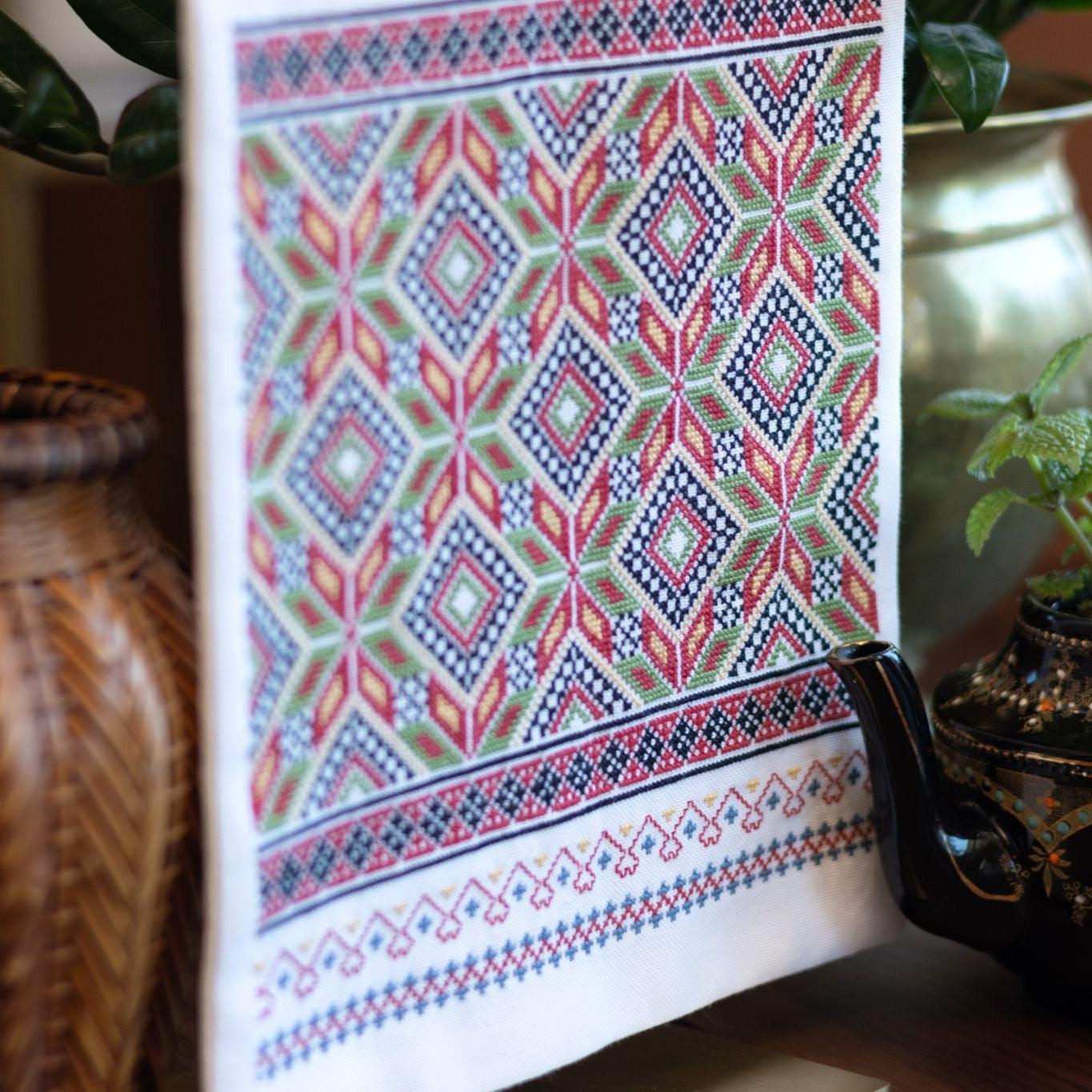 Cross stitch folk embroidery wall hanging with a colorful geometric pattern on a surface with decorative pots and plants.