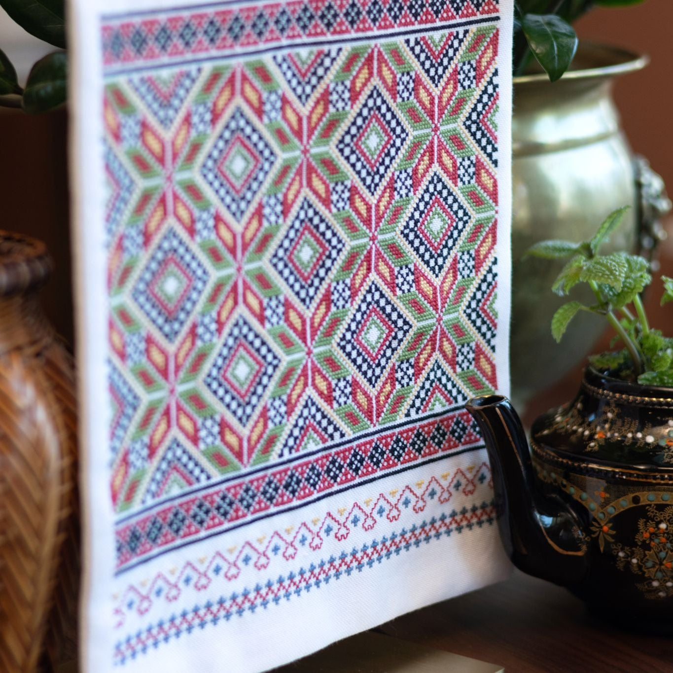 Cross stitch folk embroidery wall hanging with a colorful geometric pattern on a surface with decorative pots and plants.