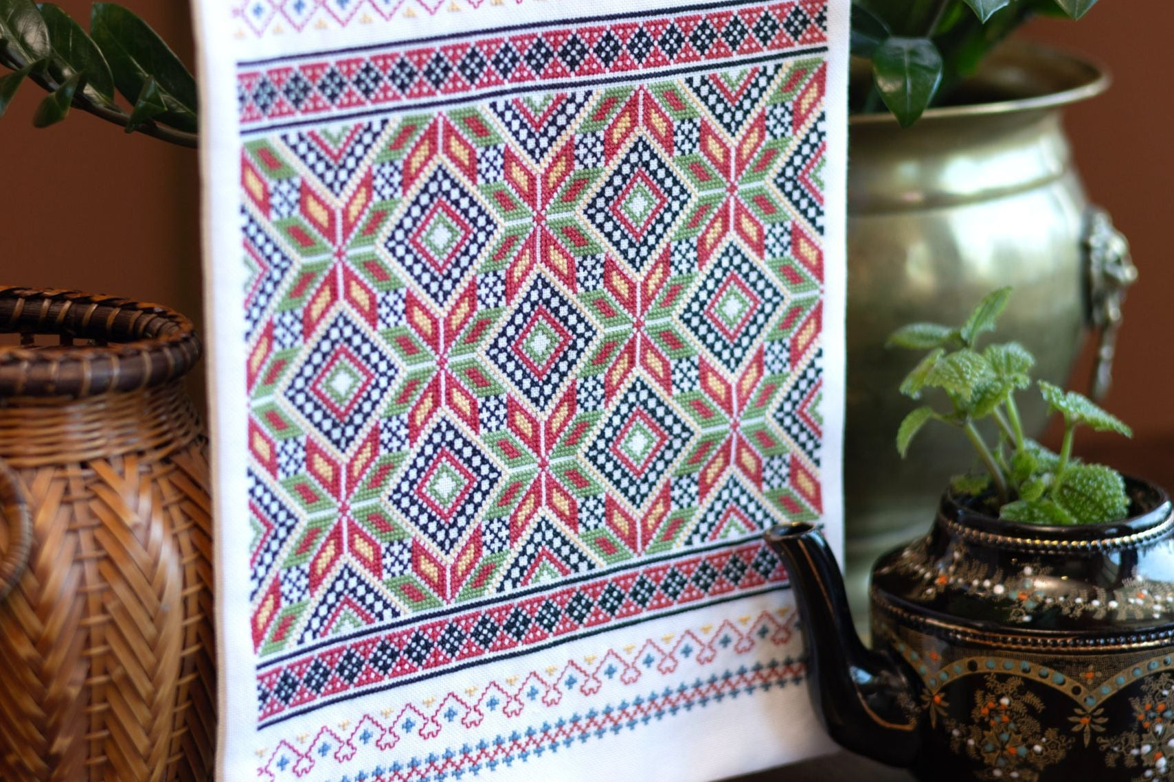 Cross stitch folk embroidery wall hanging with geometric pattern on a surface with decorative vases and plants.