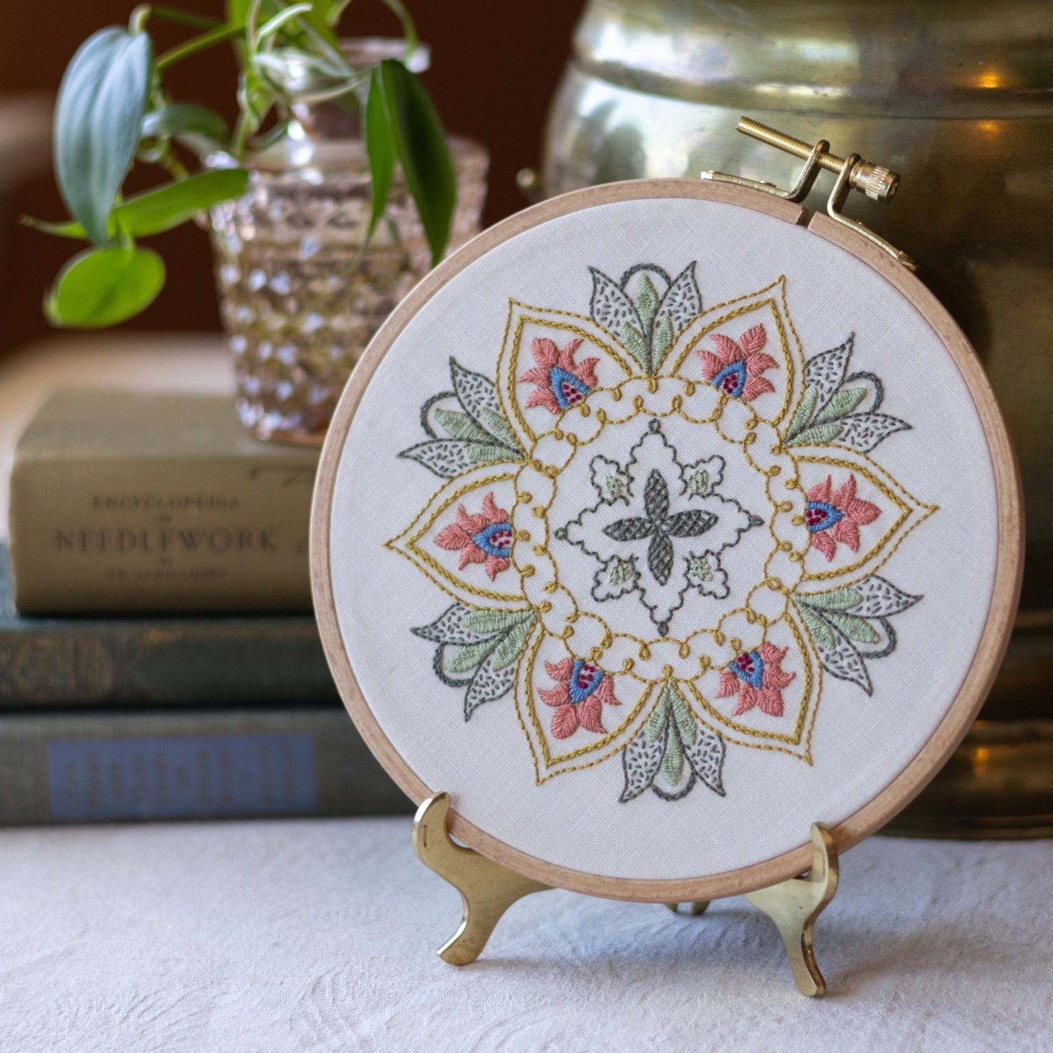 Embroidery hoop with floral design on a stand, placed on a surface with books and a plant in the background.
