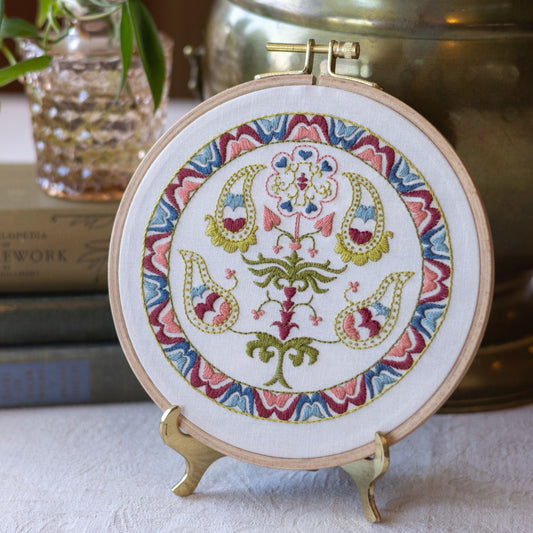 Embroidery hoop with colorful floral design on a stand, with books and a plant in the background.