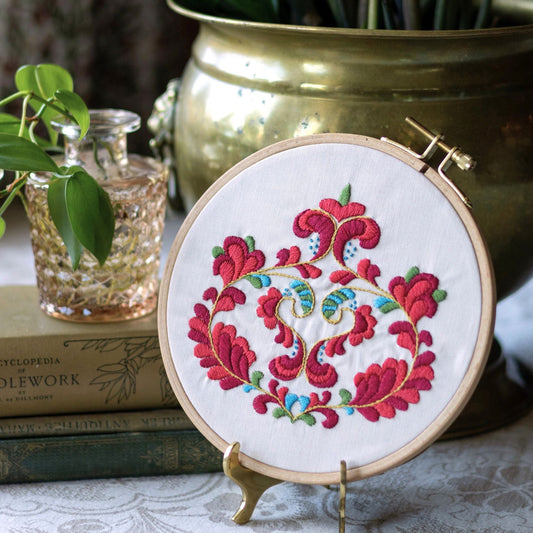Embroidery hoop with floral design on a stand, placed on a surface with books and a plant in the background.