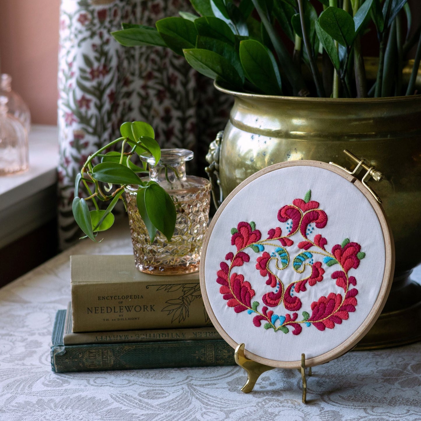 Embroidery hoop with floral design on a table with books and plants