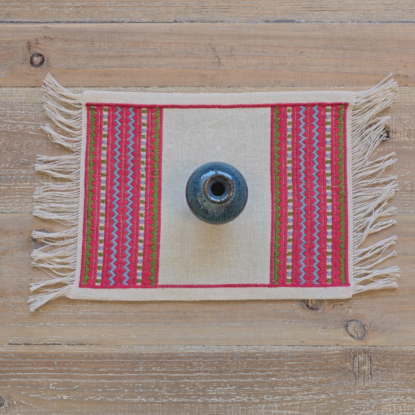 A video of an atzolati cross stitch folk embroidery textile with fringe, featuring a red border and geometric stripes in light blue and green, styled on a wooden table with a blue ceramic vase.